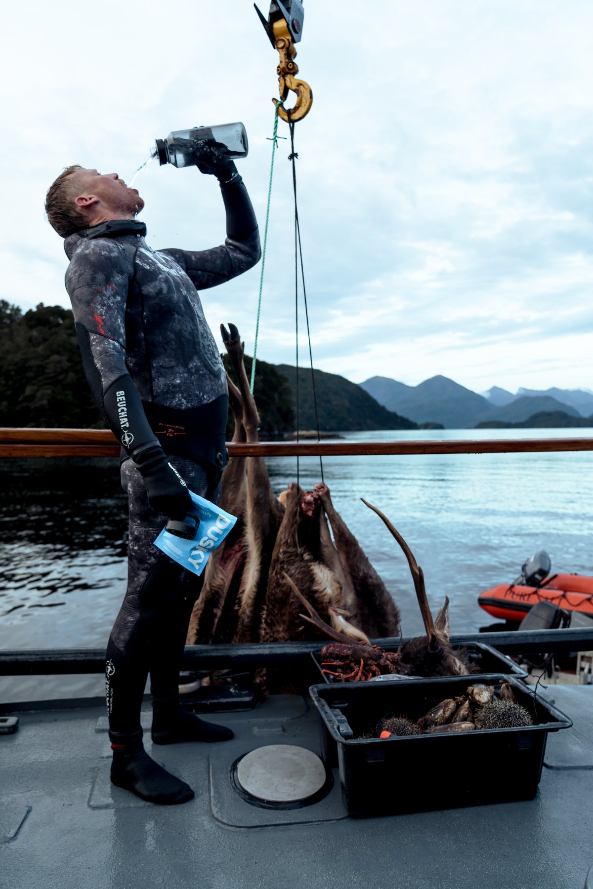 Spearfisherman having Dusky electrolyte capsules with water bottle aboard a boat after a successful day in Fiordland, with deer, fish and sea urchins on deck and mountains in the background.