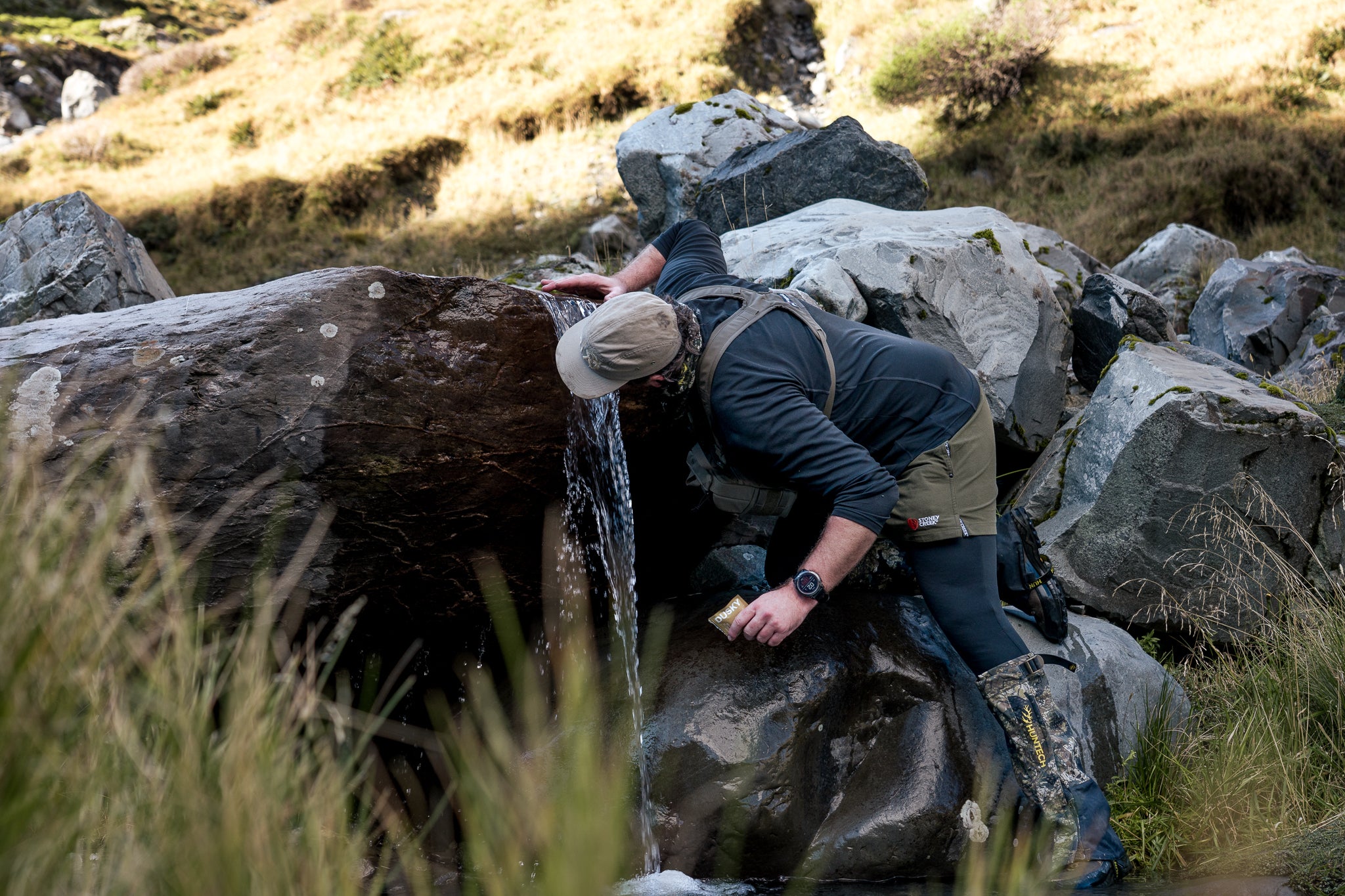 Hunter illing a water bottle from a mountain stream while carrying Dusky electrolyte and energy capsules in New Zealand. 