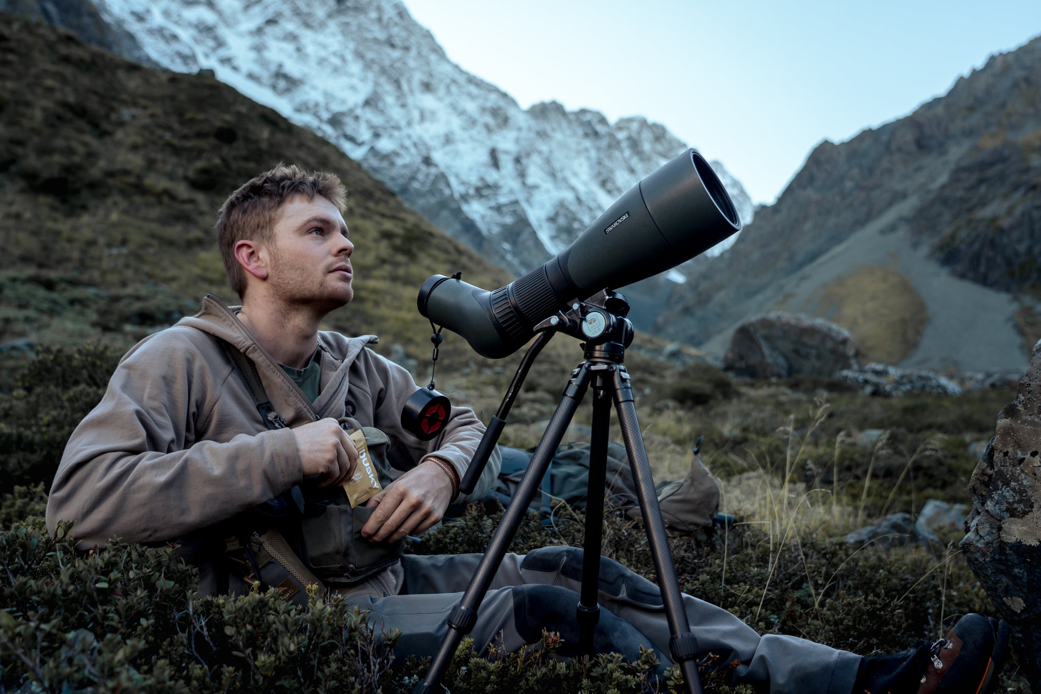 Hunter glassing alpine terrain with a spotting scope while carrying Dusky electrolyte and energy capsules during a backcountry trip.